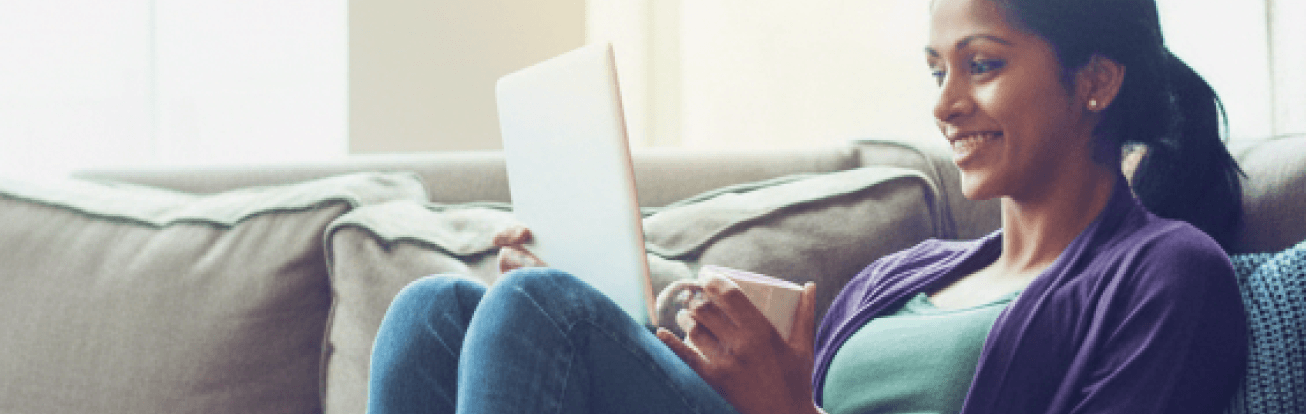 A causally dressed woman smiles at her laptop screen while holding a cup of coffee leaning back against her sofa.